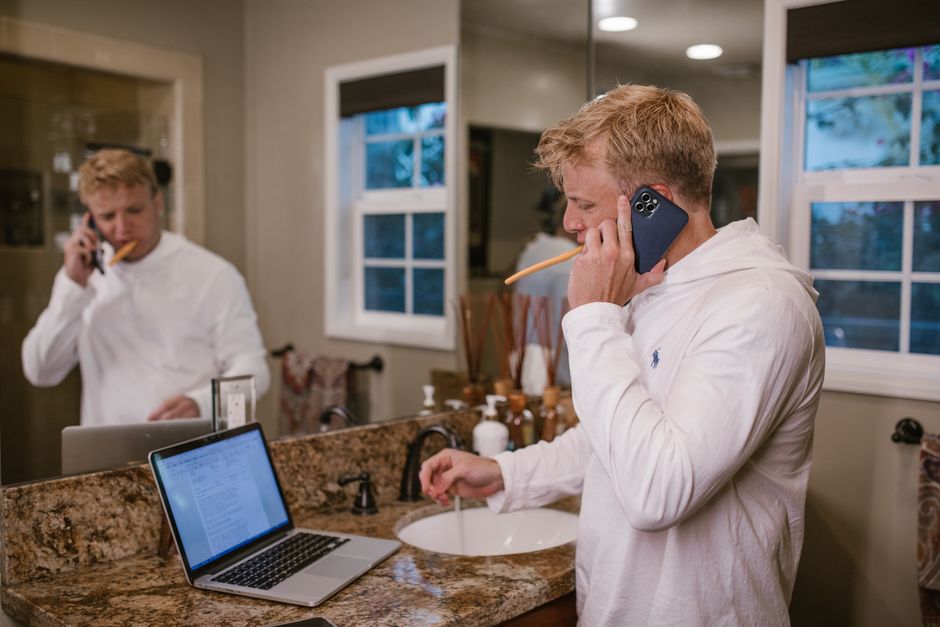 Man brushing teeth and on phone in bathroom, multitasking over laptop.